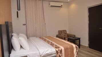 Quarto de hotel com uma cama, uma cadeira, uma mesinha, ar condicionado e uma porta.