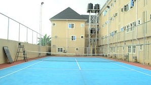 An indoor tennis court with a net, surrounded by a metal fence and a building with multiple windows and air conditioning units. - Newton Hotels Limited Owerri (Owerri)