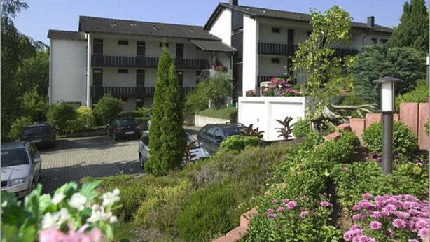Un hotel de varios pisos con estacionamiento al frente, rodeado de follaje y flores.