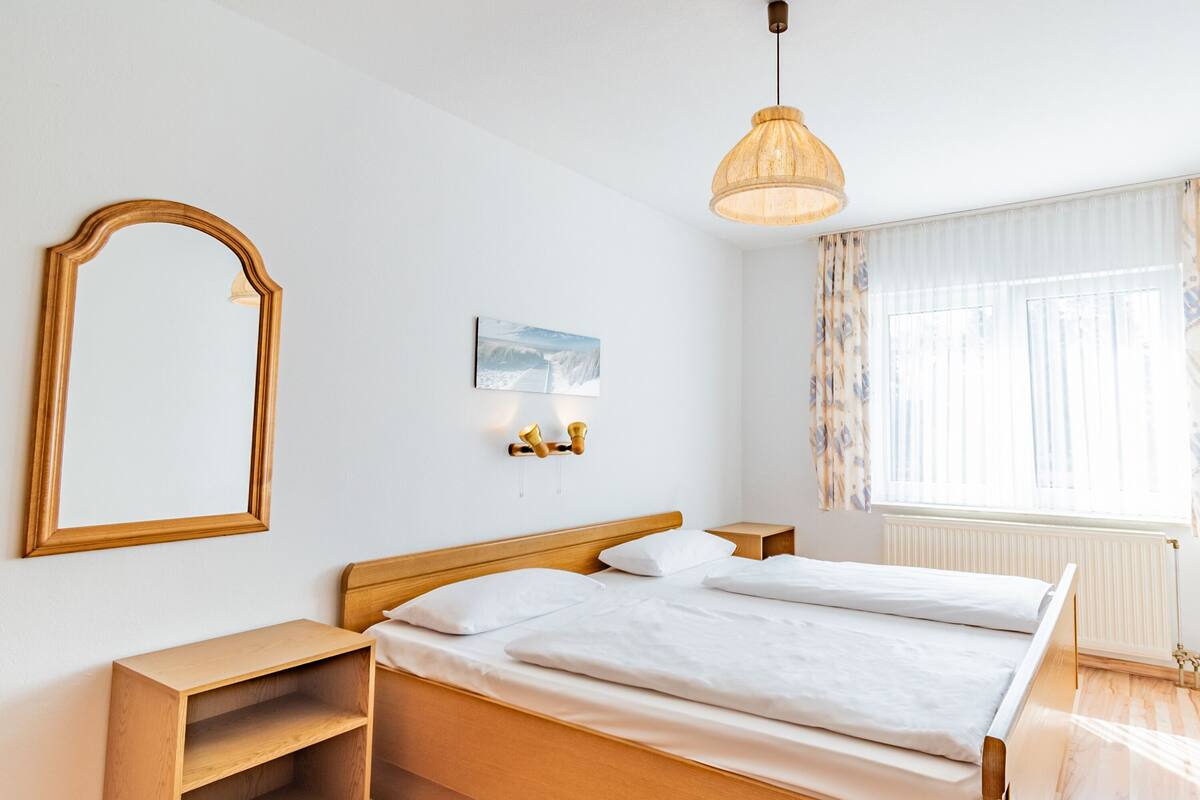 A neatly arranged bedroom with a wooden bed, a bedside table, a mirror, and a hanging light fixture.