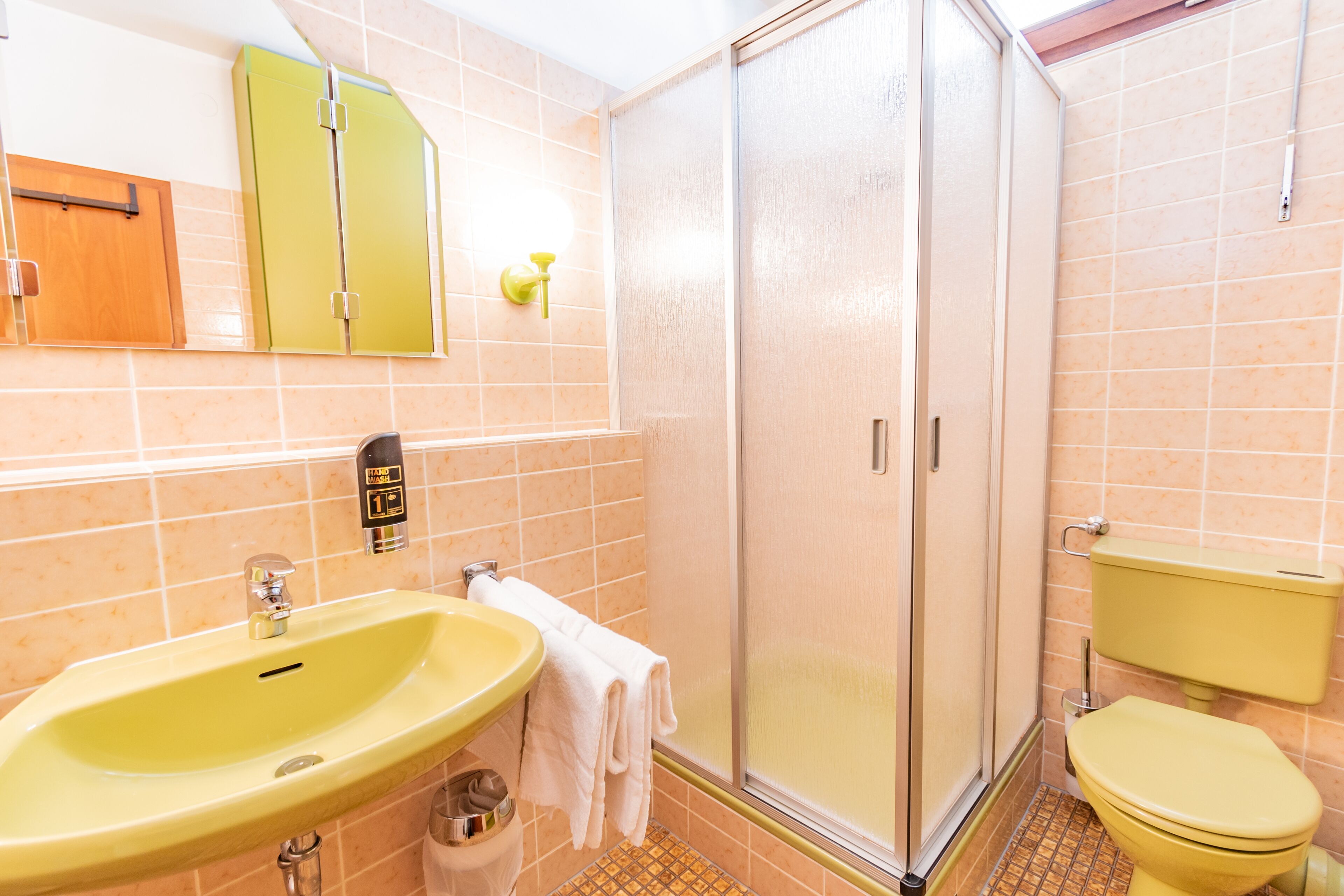 Un baño con un lavamanos amarillo, un inodoro, una cabina de ducha y un espejo con un armario verde encima.