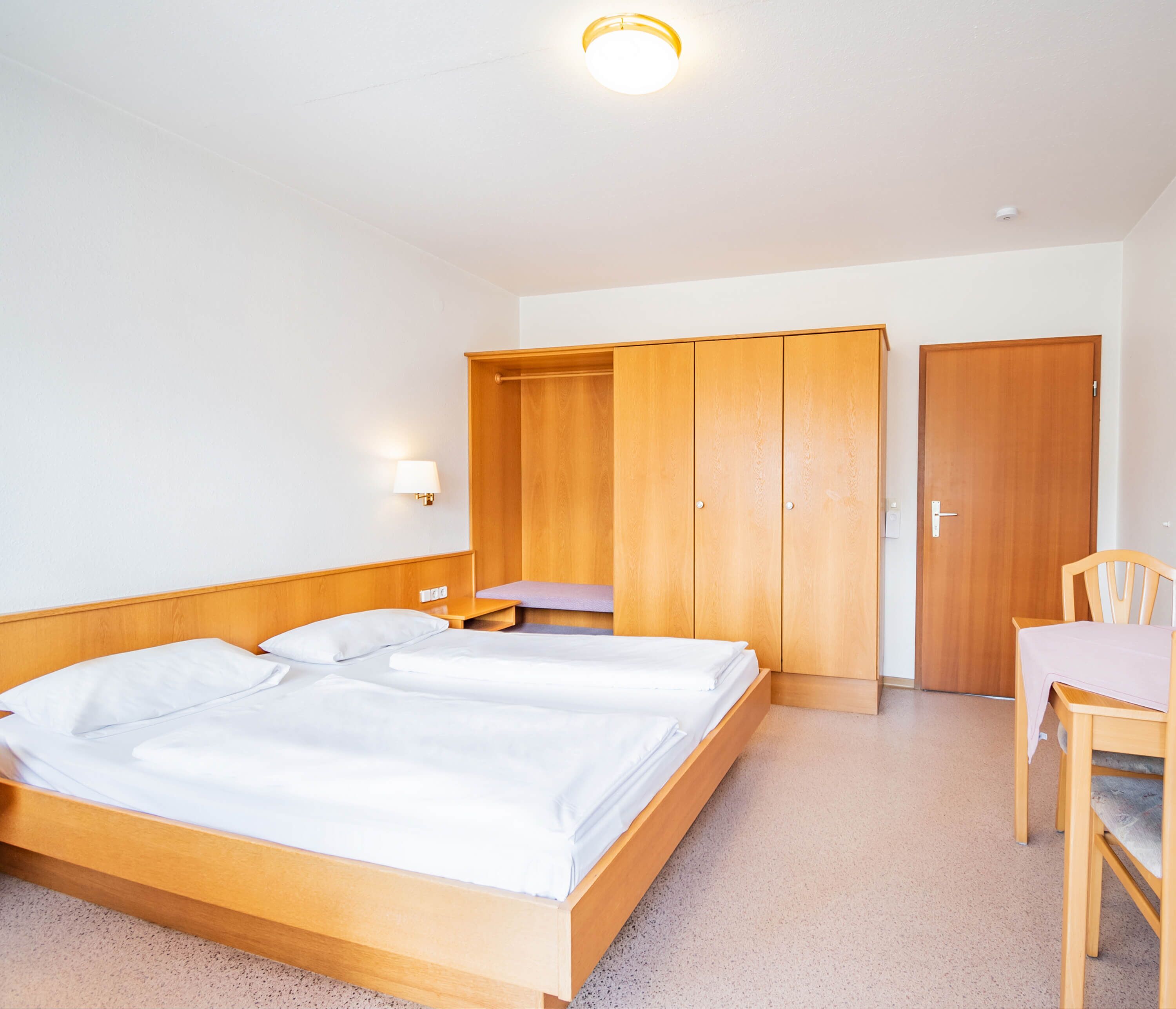 A bedroom with a double bed, wooden wardrobe, a desk with a chair, and a wall-mounted lamp.