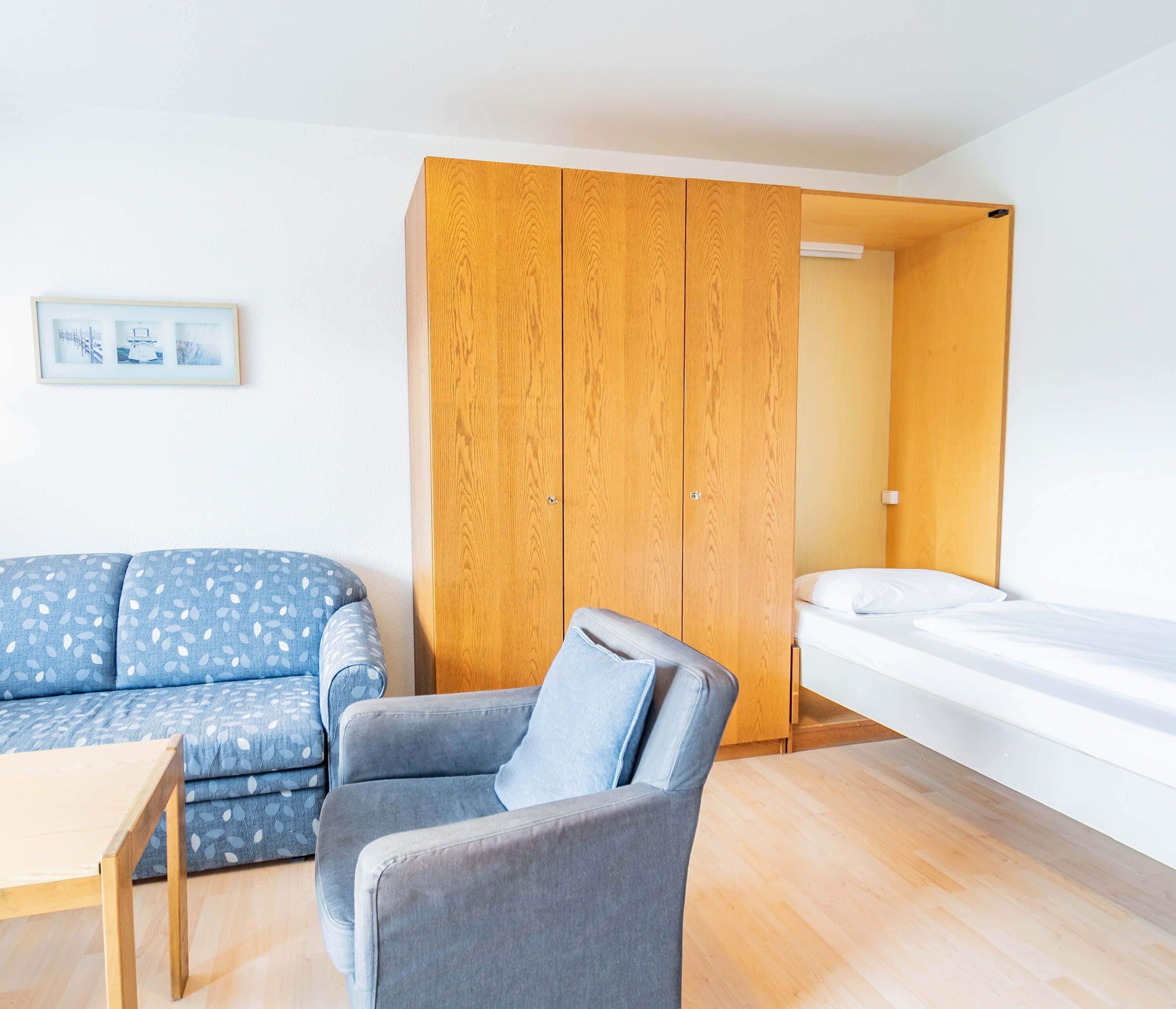 A modern, compact room with a wooden wardrobe, a grey armchair, a blue sofa, and a bed.