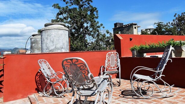A rooftop terrace with two metal rocking chairs and a table, surrounded by a red wall and brick flooring. - Hostal Tony y Daisy TRINIDAD (Trinidad)