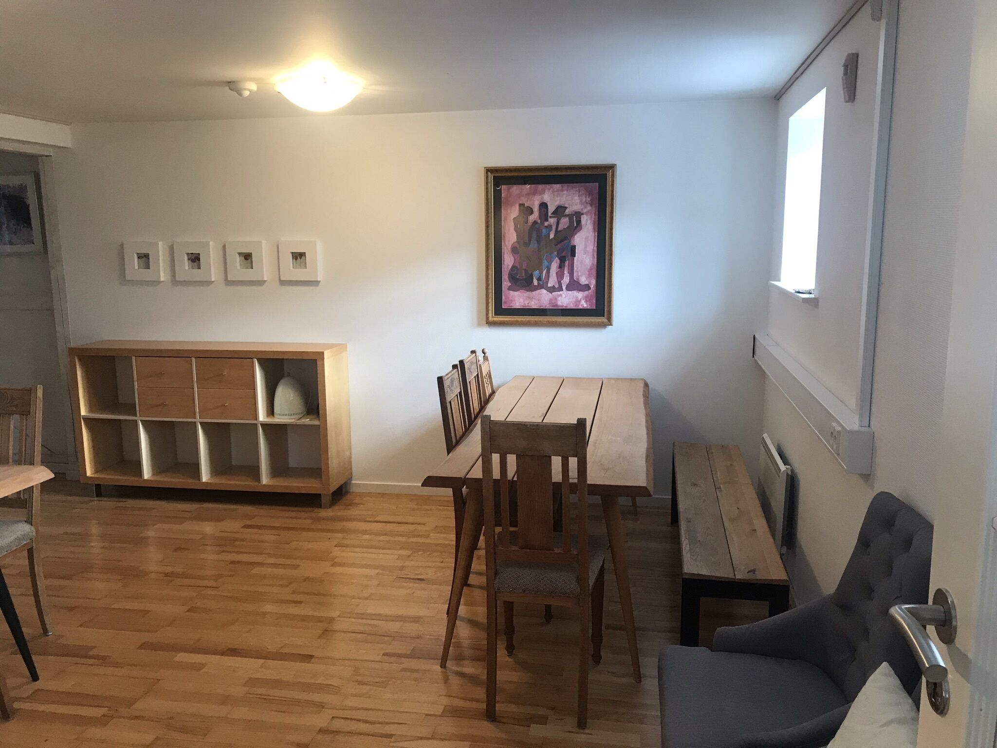 A room with a wooden floor, a dining table with chairs, a cushioned armchair, a wooden cabinet, and a framed artwork on the wall.