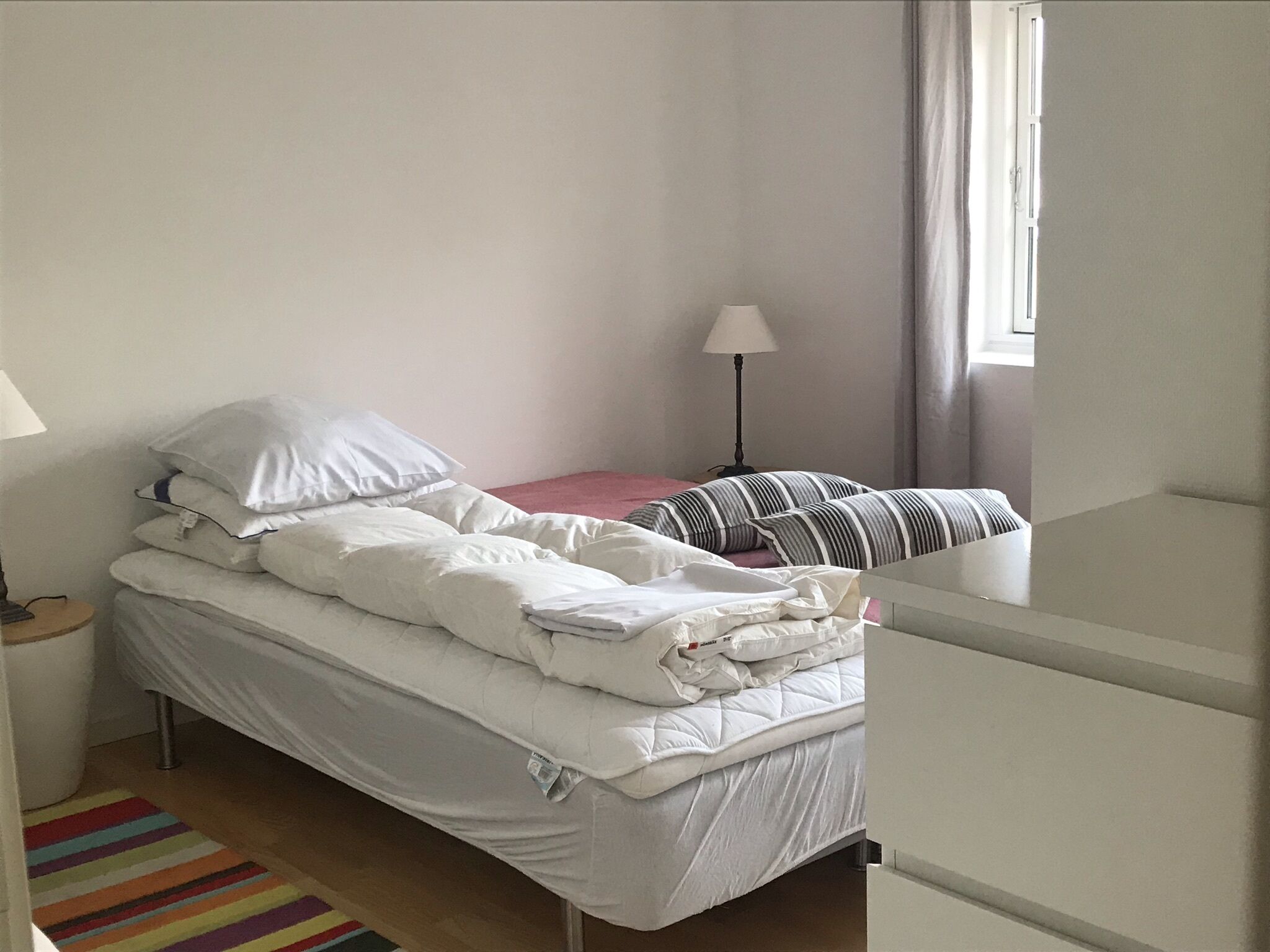 A single-bed room with a white bed, a bedside table, a lamp, a striped pillow, and a window with curtains.