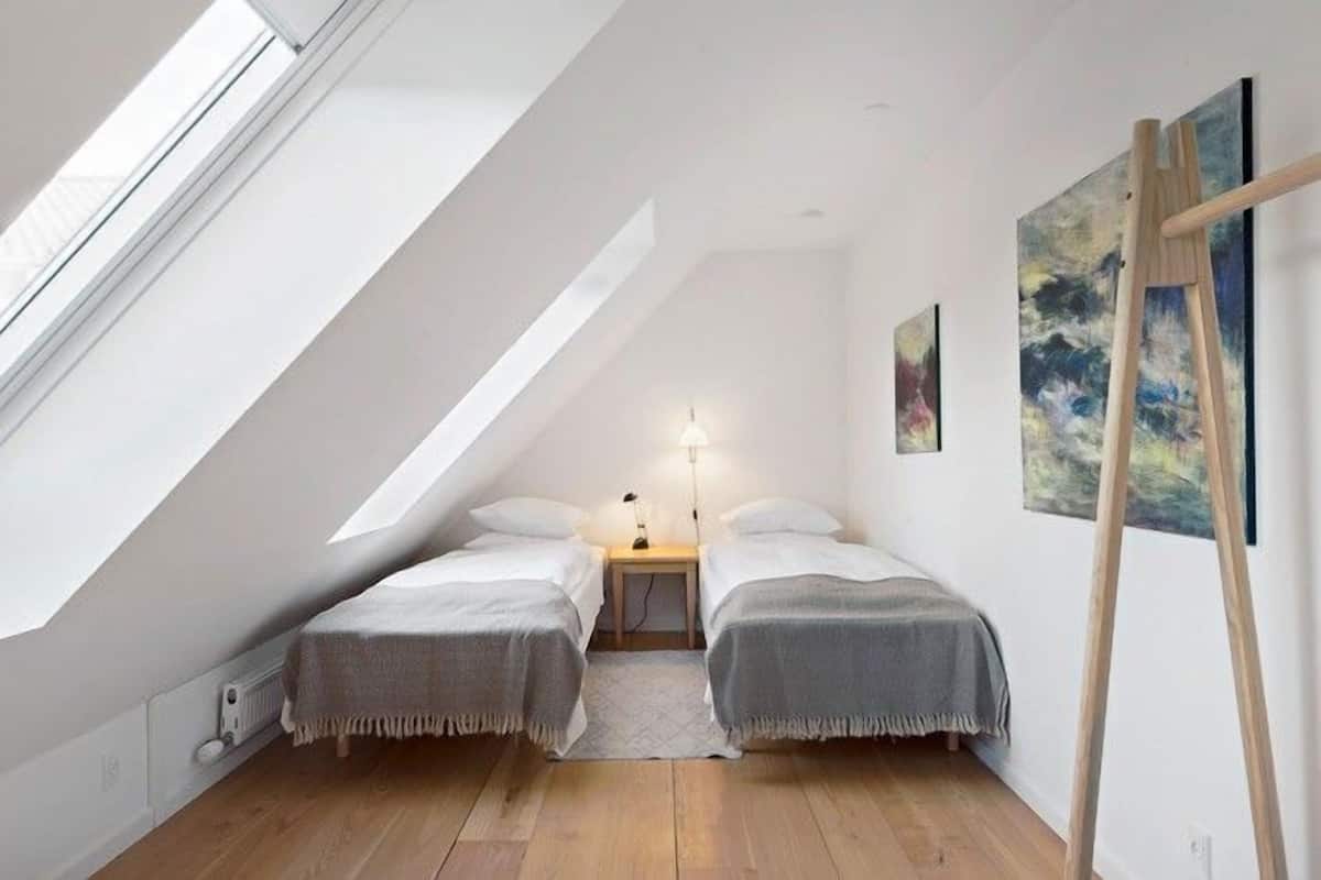A small, cozy attic bedroom with two beds, a skylight, a wooden ladder, and a painting on the wall.