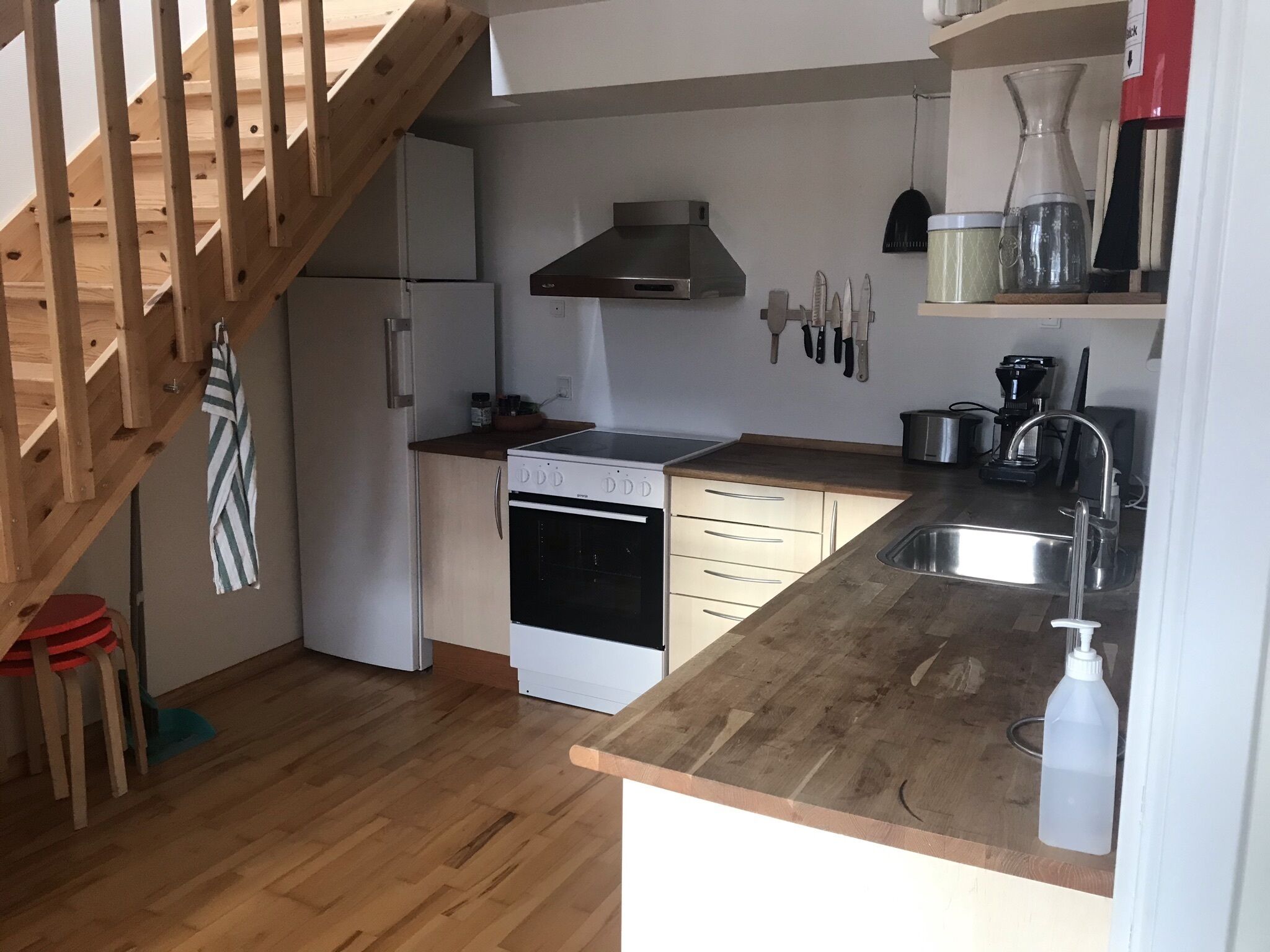A compact kitchen with a stove, oven, and sink. There is a wooden staircase on the left, and a shelf with kitchen utensils above the counter.