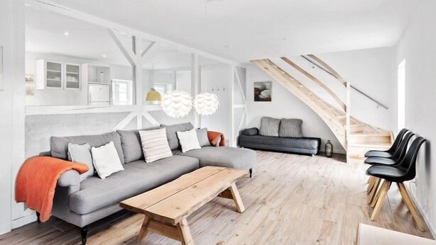 Una sala de estar moderna con un sofá seccional gris, una mesa de centro de madera y sillas negras. Una escalera con barandillas de madera conduce a un nivel superior.
