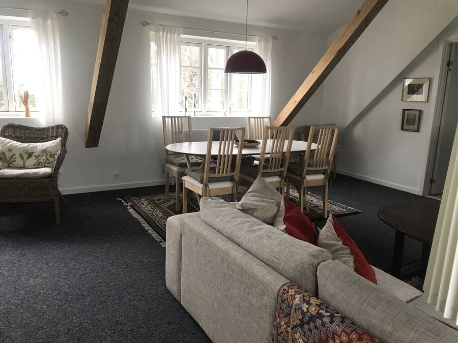 Una sala de estar con un sofá, una mesa de comedor con sillas, una lámpara colgante y una ventana con cortinas.