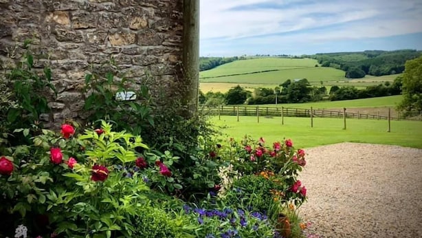 A stone wall with a sign for Sproxton Hall Holiday Cottages, surrounded by colorful flowers and a view of green fields and hills.
