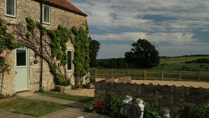 A stone house with a green door and windows, a garden with a table and chairs, and a view of the countryside.