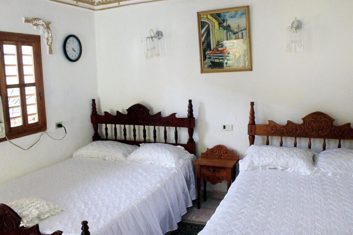 A bedroom with two wooden beds, a small bedside table, a wall clock, a painting, and a window with blinds.