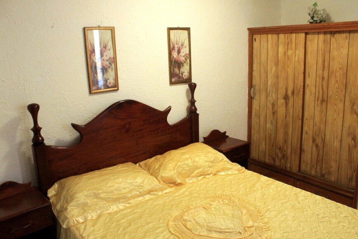 A bedroom with a wooden bed, a yellow bedspread, two framed floral pictures on the wall, and a wooden wardrobe.