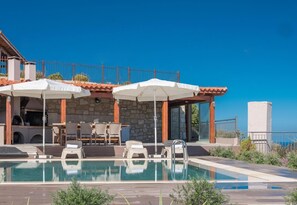 A pool area with lounge chairs and umbrellas, a stone wall, and a view of the sea. - Eolos (Malevizi)