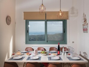 A dining area with a table set for four, a view through a window, and a clock on the wall. - Eolos (Malevizi)