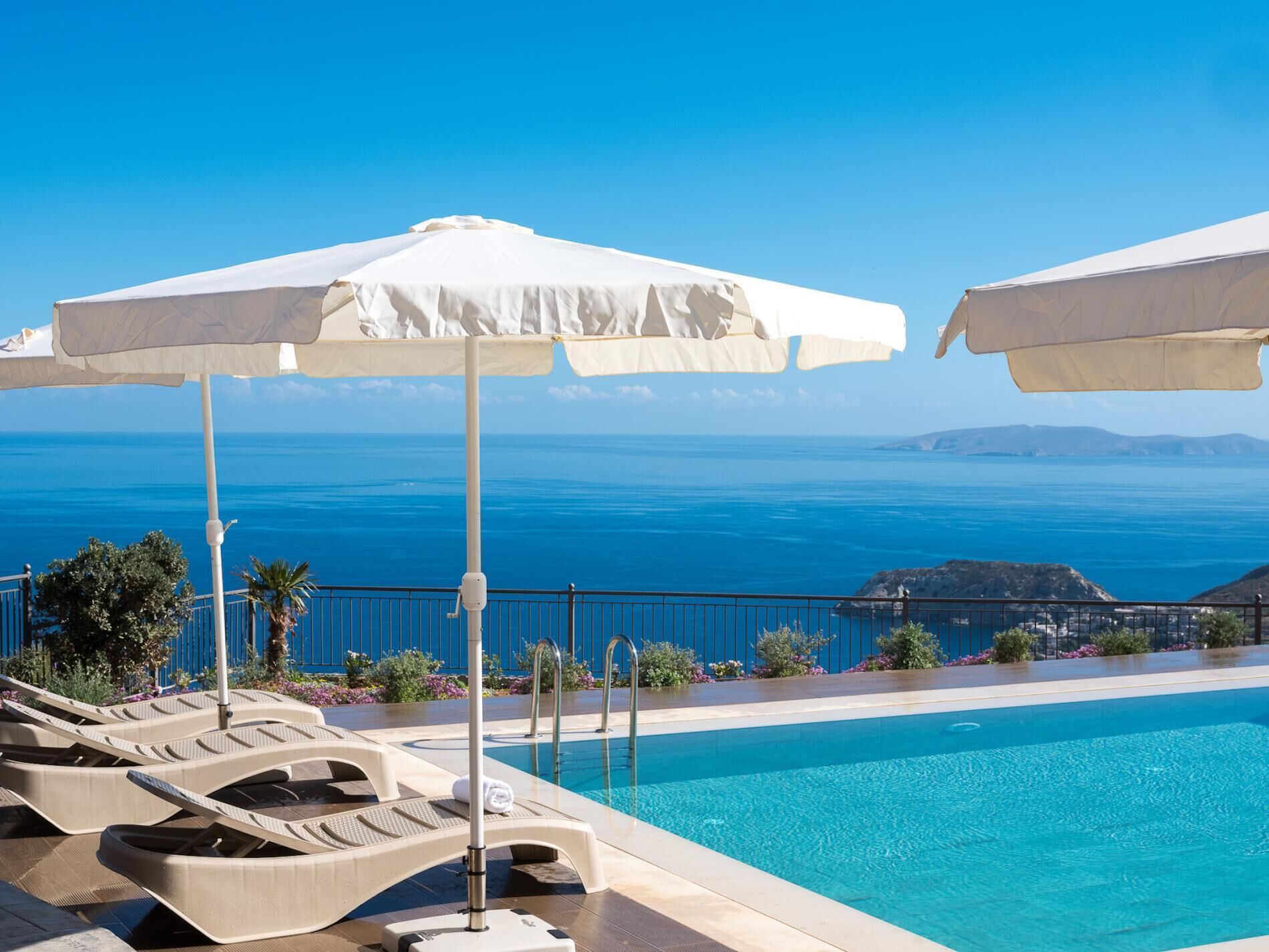 A poolside area with lounge chairs and umbrellas, overlooking a clear blue sea and distant land.
