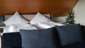 Quarto com teto de madeira, cama com almofadas brancas e azul-escuras, luminária de cabeceira e planta em vaso.