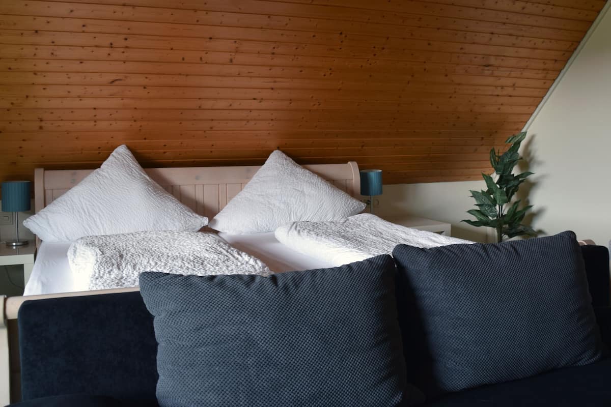 A bedroom with a wooden ceiling, a bed with white and dark blue pillows, a bedside lamp, and a potted plant.