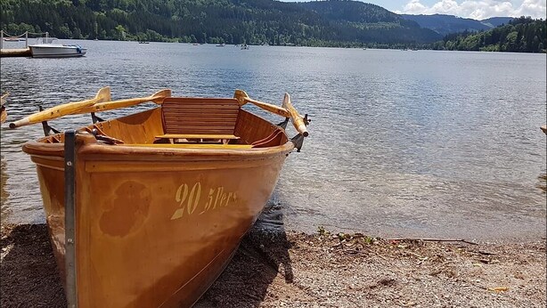 Um barco de madeira com o número 20 na lateral, atracado em um lago com montanhas arborizadas ao fundo.
