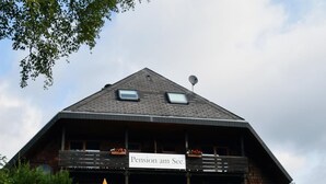 Um edifício de madeira de dois andares com varandas, uma placa com a inscrição 'Pension am See' e área de estar ao ar livre com guarda-sóis.