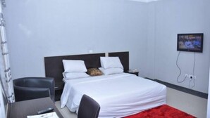 A hotel room with a bed, a desk, a chair, a TV, and a red carpet. - Mac View Hotels (Owerri)