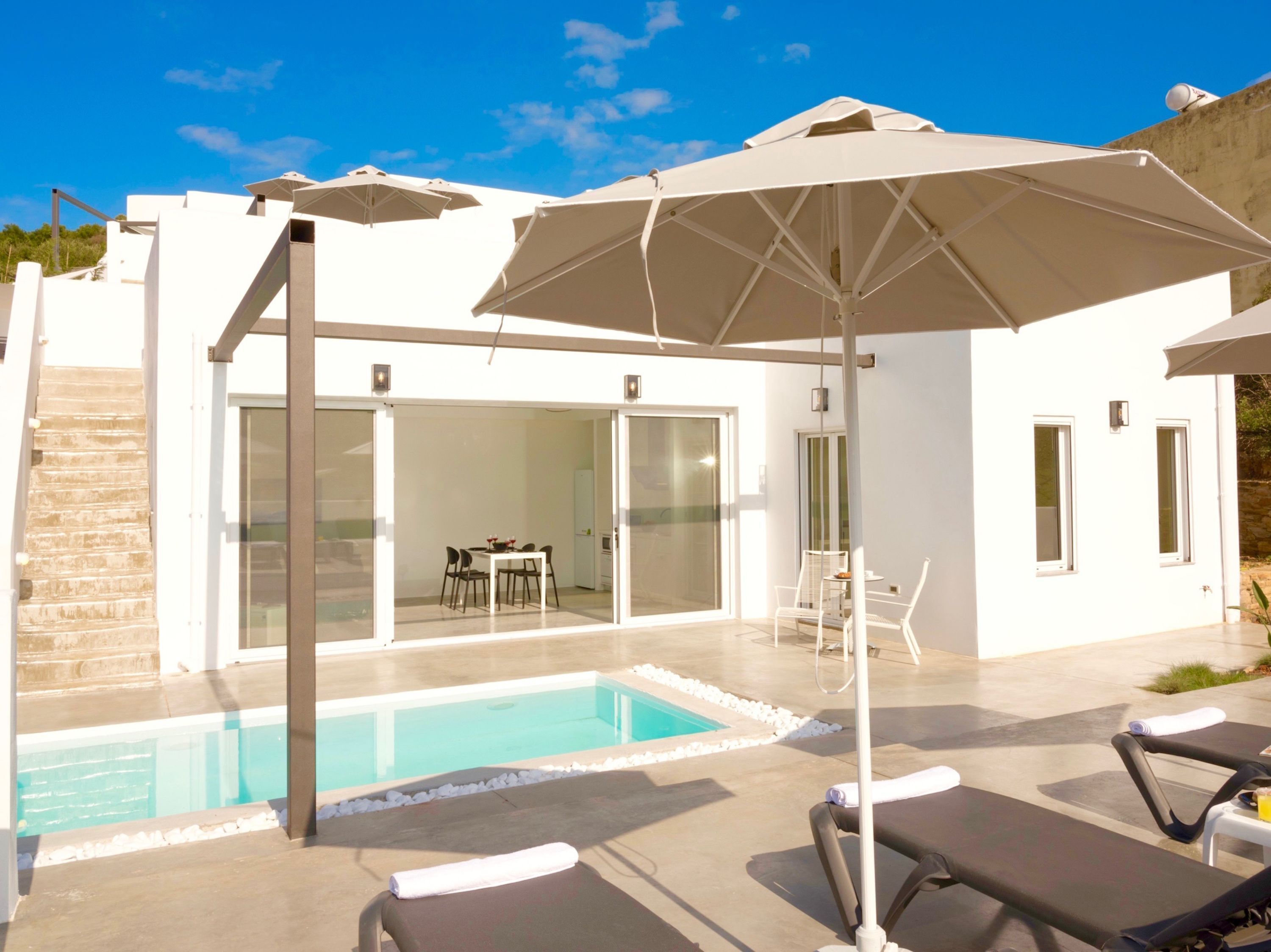A modern pool area with lounge chairs and umbrellas, a dining area with a table and chairs, and a glass door leading to an interior space.