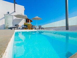 A clear blue pool with lounge chairs and umbrellas, a stone wall, and a white building with a balcony. - Paradiso I (Malevizi)
