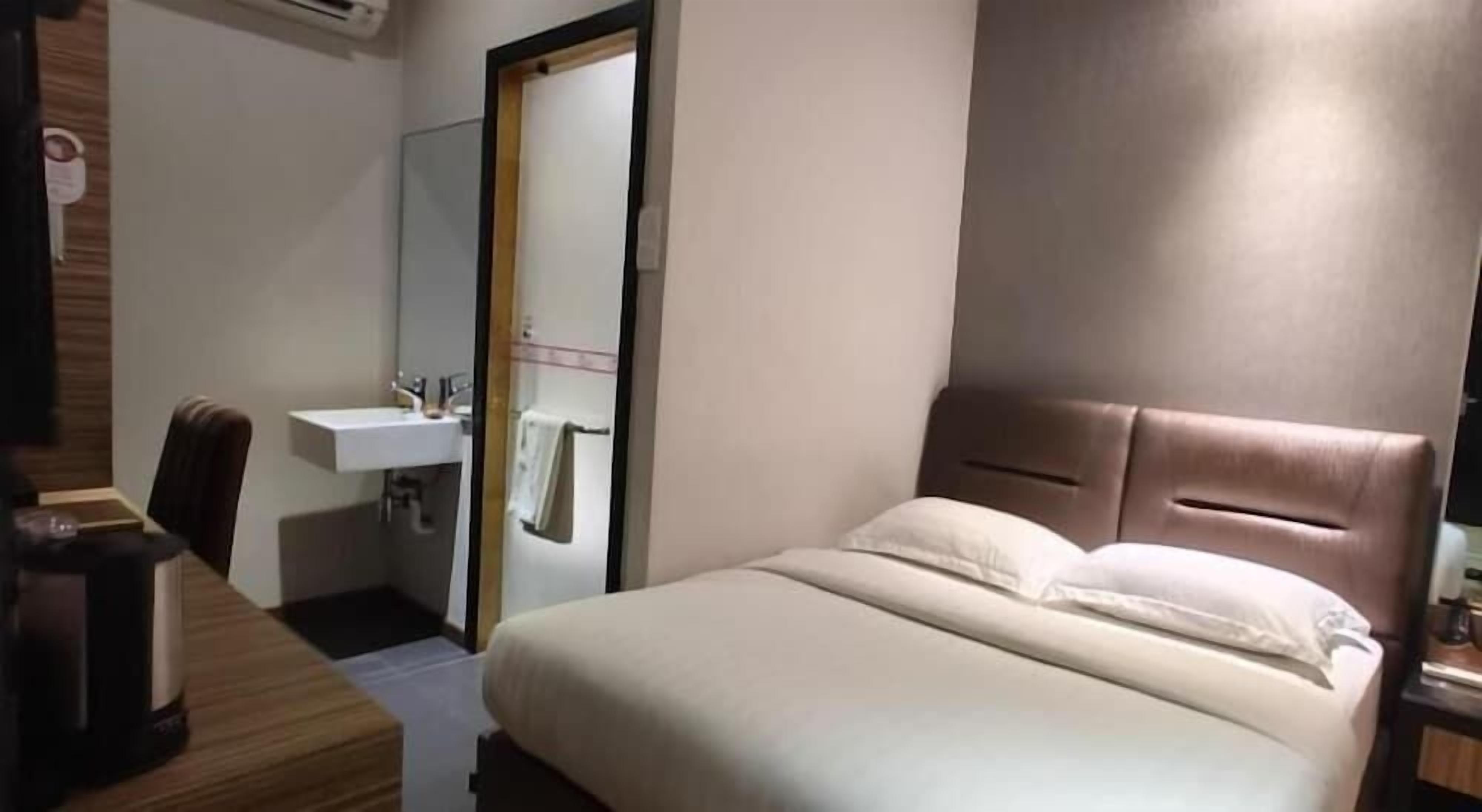 A hotel room with a bed, a bedside table, a chair, a desk, and a bathroom with a sink and mirror.