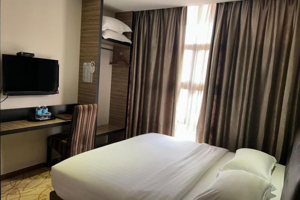 A hotel room with a bed, a TV mounted on the wall, a desk with a chair, and a window with curtains.