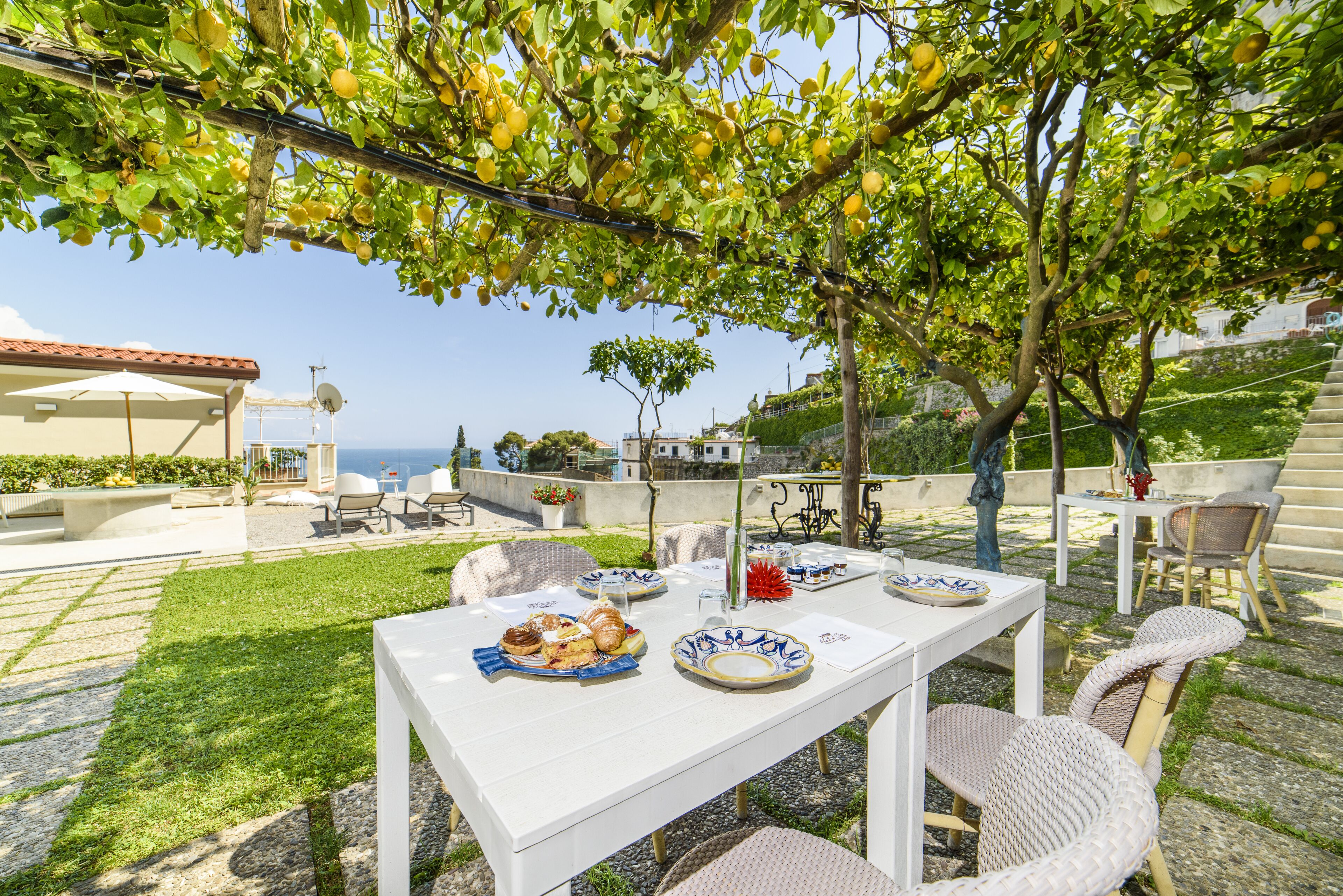 Un espace de restauration en plein air avec une table dressée pour deux personnes, entouré de chaises en osier, sous un arbre chargé de citrons mûrs.