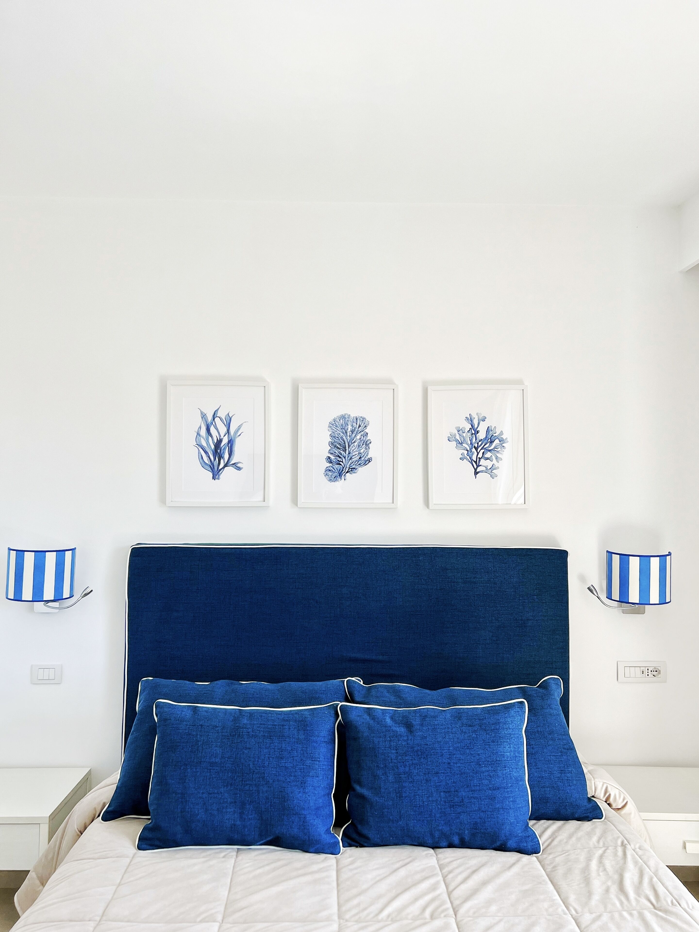 A bedroom with a blue headboard, white bedding, and three framed botanical prints on the wall.