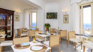 Uma sala de jantar com mesas e cadeiras brancas, um armário de madeira e vista para o mar.