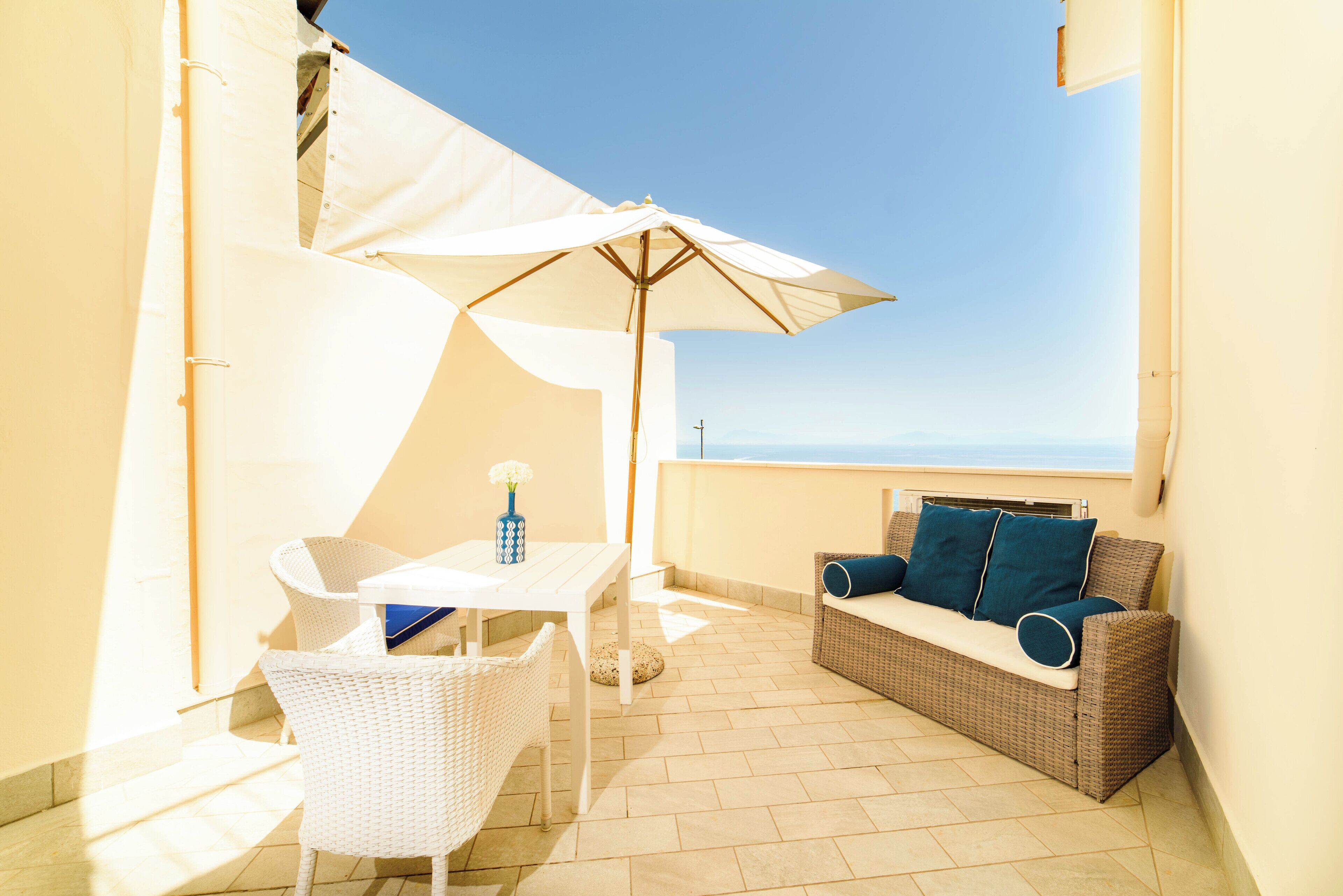 A balcony with a white table, wicker chairs, a wicker sofa with cushions, and a large umbrella.