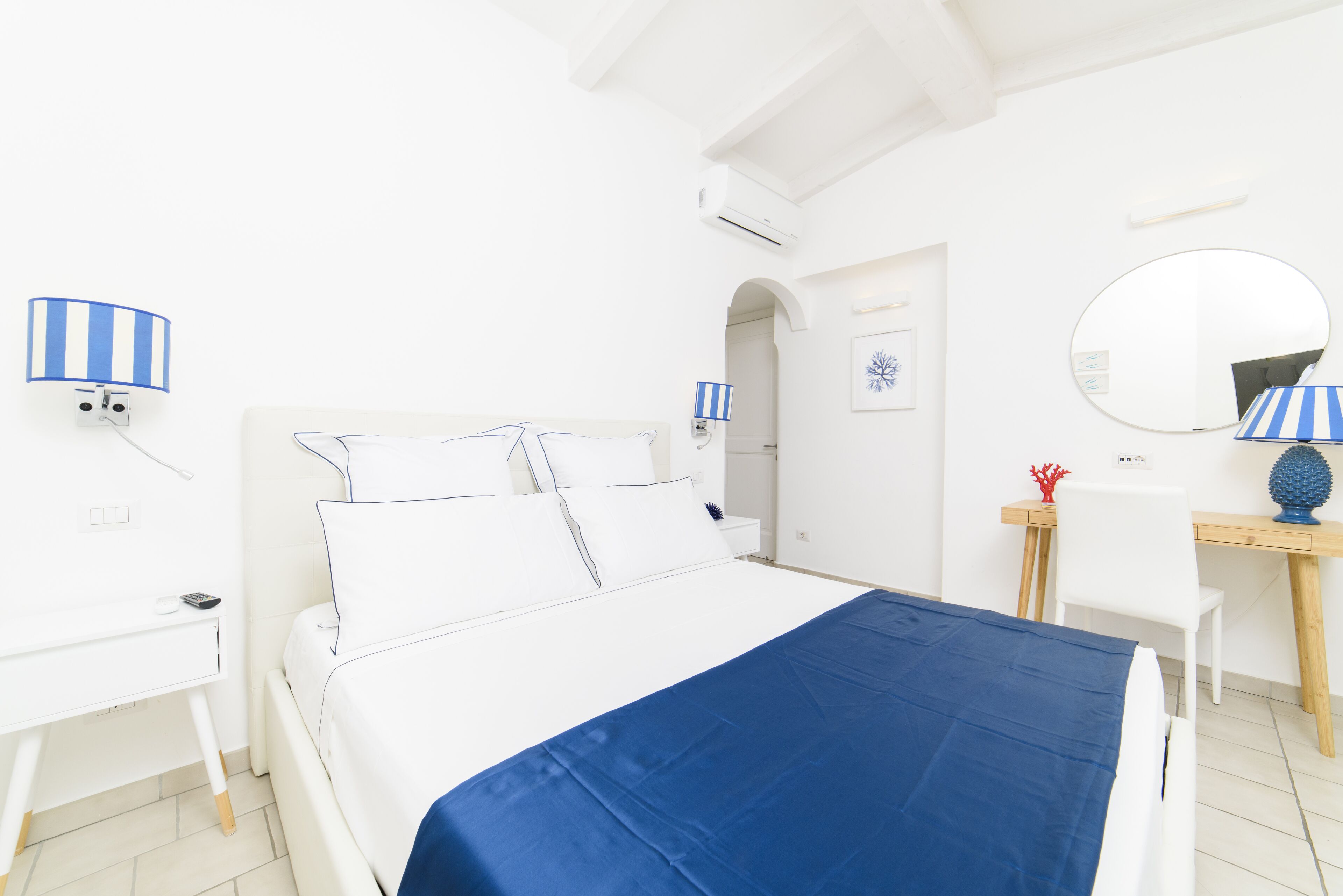 Une pièce blanche avec un lit, un bureau et une chaise. Il y a un miroir au mur et une lampe sur le bureau.