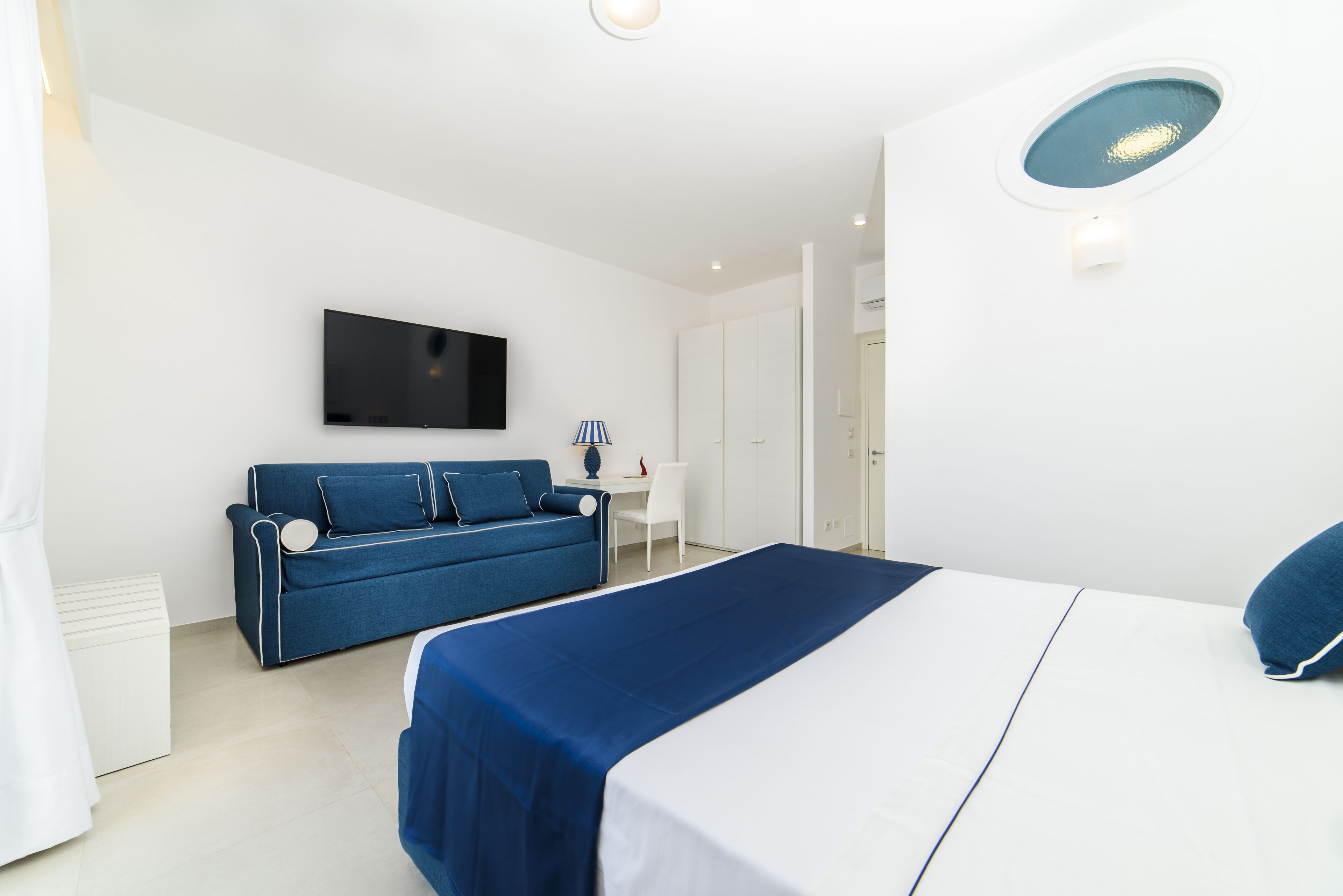 A modern hotel room with a large bed, a blue sofa, a flat-screen TV, and a small desk with a lamp.