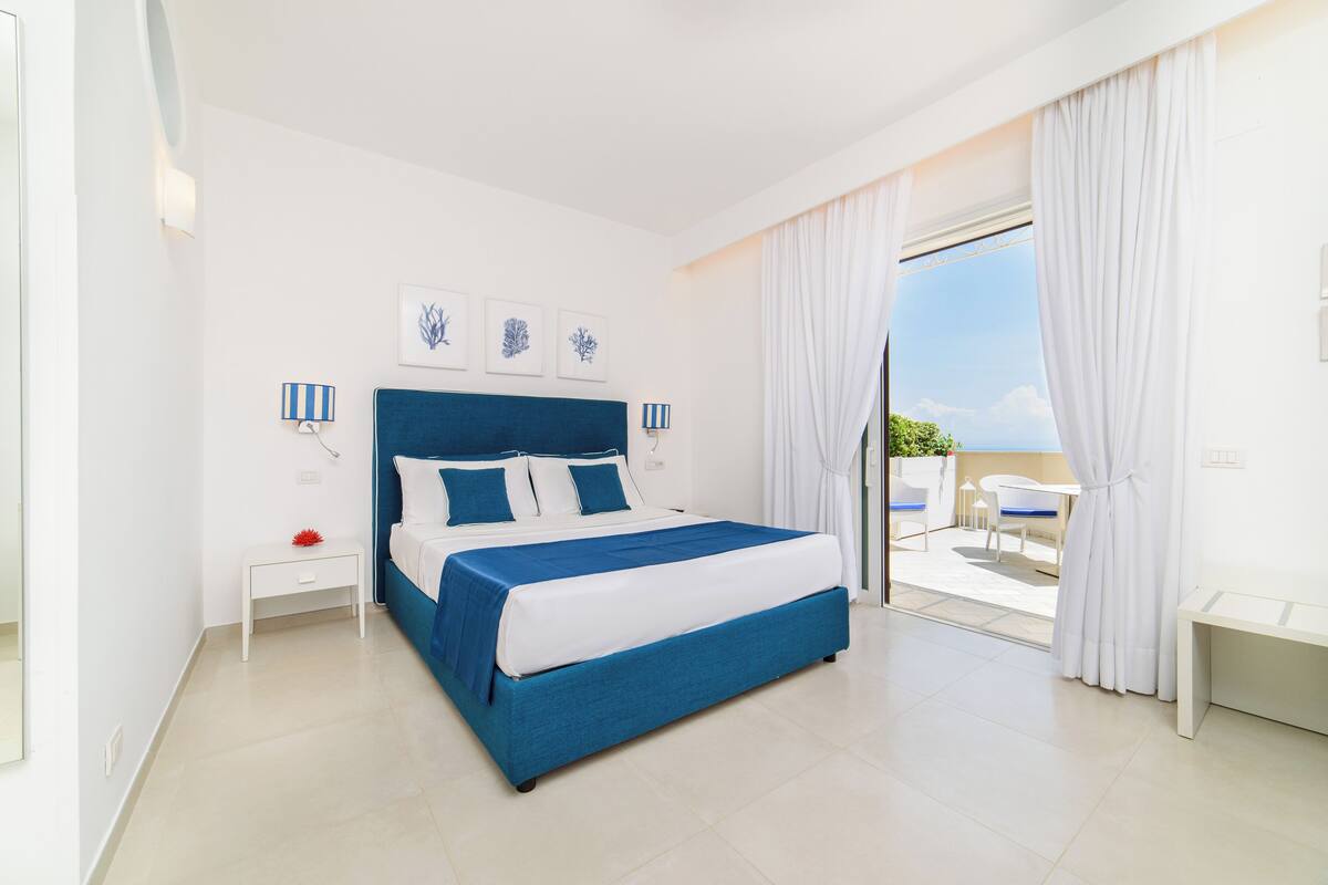 Une chambre d’hôtel avec un grand lit, une petite table de chevet, une commode, une fenêtre avec des rideaux et une vue sur l’extérieur.