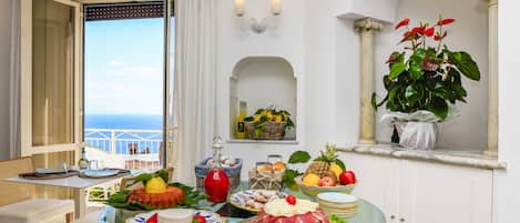 A dining table with a variety of food items, a view of the sea through an open window, and a potted plant on a shelf.