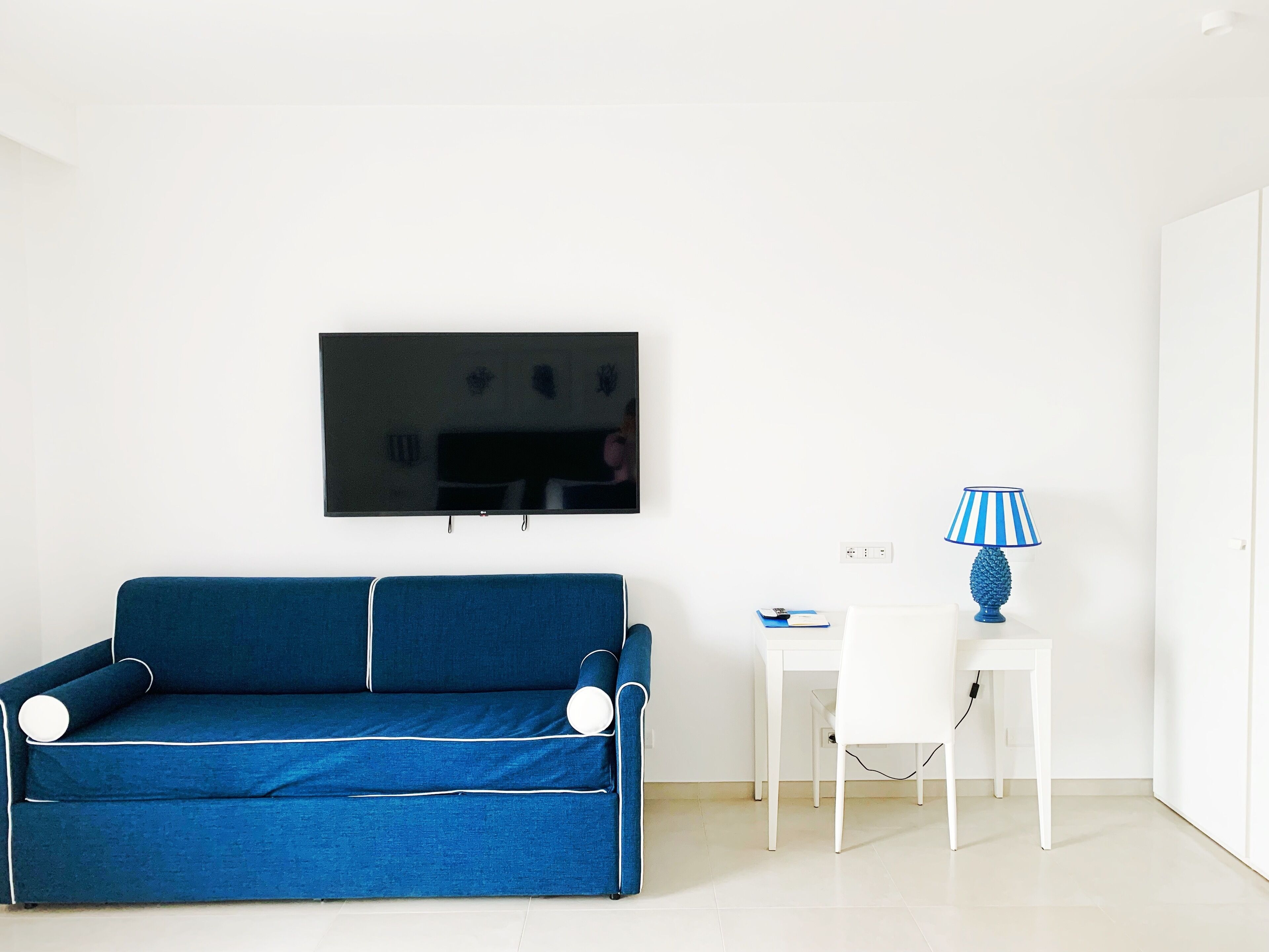 Ein Zimmer mit einem blauen Sofa, einem weißen Schreibtisch, einer Lampe und einem Fernseher an der Wand.