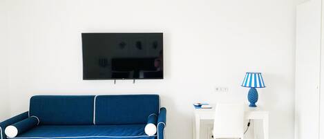 A room with a blue sofa, a white desk, a lamp, and a TV on the wall.