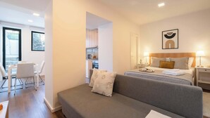 A modern hotel room with a sofa, a bed with a headboard, a dining area with a table and chairs, and a kitchen in the background.