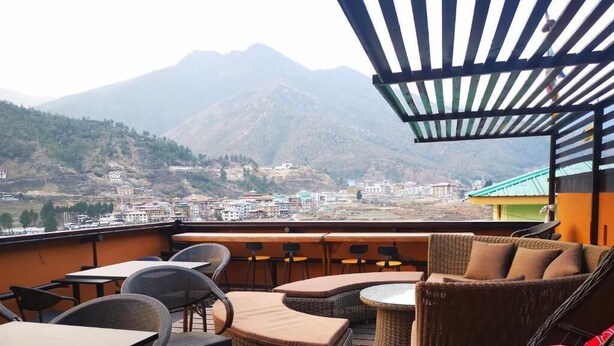 Una terraza con mobiliario de mimbre, vistas a un paisaje montañoso y un edificio al fondo.