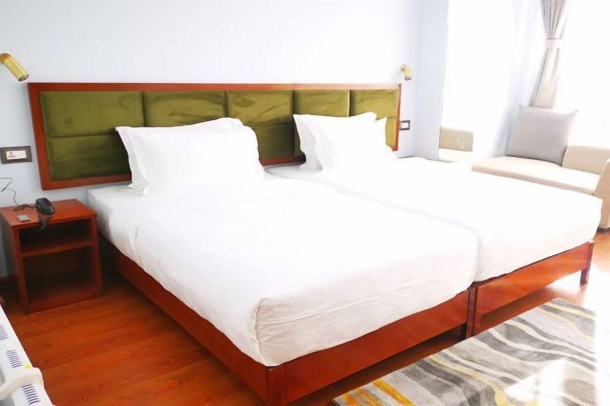 A double bed with a wooden frame and green headboard, two bedside tables, a chair, and a window with curtains.