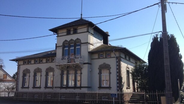A two-story building with a symmetrical facade, a central entrance with a decorative canopy, and a small balcony above the main entrance. - L' Obsidienne (Lixheim)