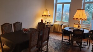 A dining area with a table and chairs, a lamp, a radiator, and a window with a view of trees. - L' Obsidienne (Lixheim)