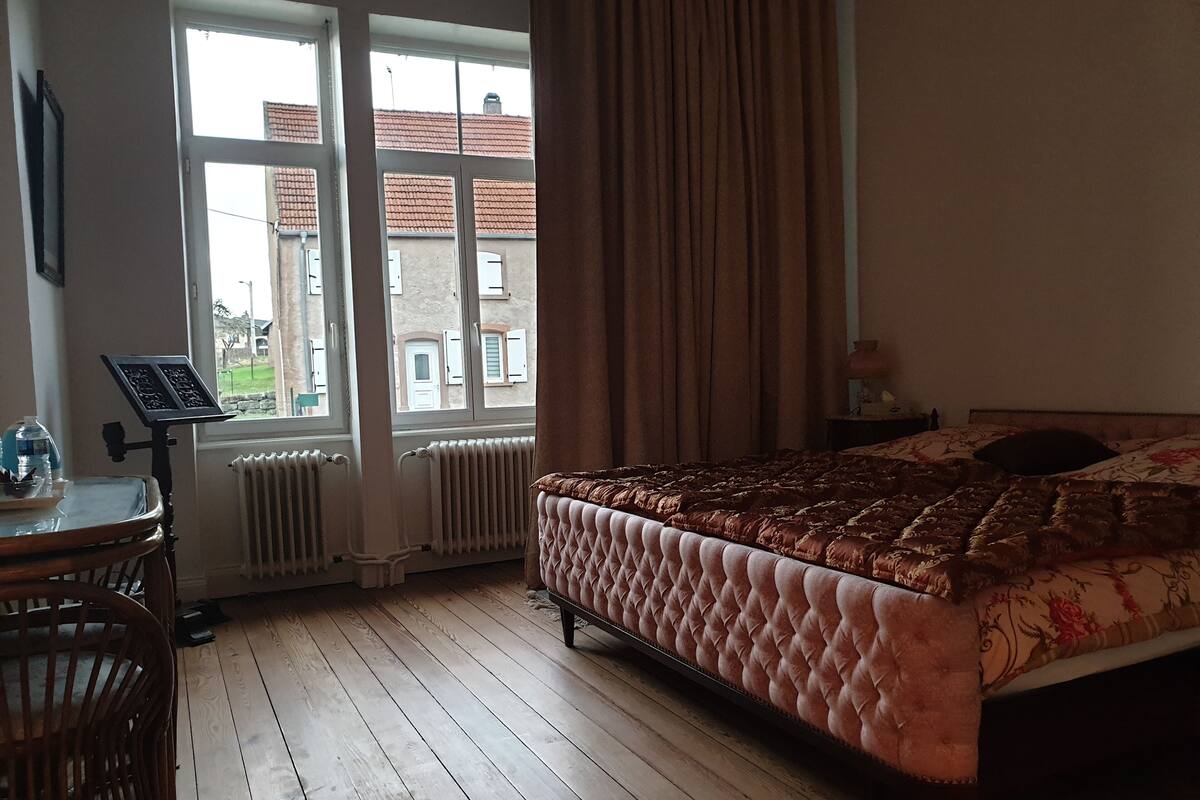 A bedroom with a large bed, wooden floor, a radiator, a window with curtains, and a small table with a music stand.
