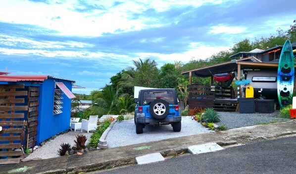 A blue vehicle parked in a driveway with a small building and a kayak in the background.