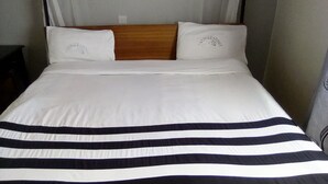 Un lit avec une literie blanche et une couverture à rayures, une tête de lit en bois et un oreiller blanc orné d’un logo.