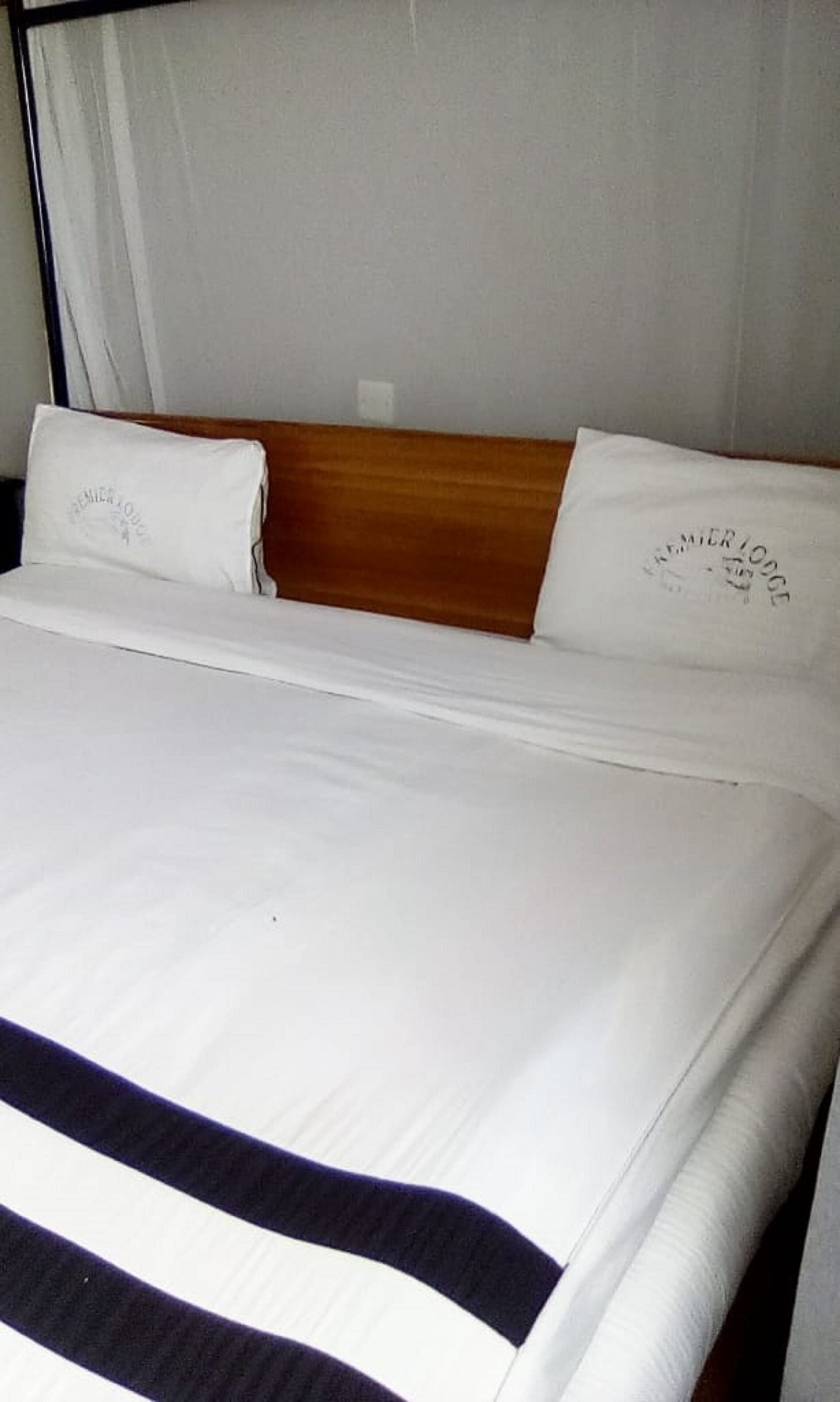 A neatly made bed with white bedding and a dark horizontal stripe, a wooden headboard, and a white curtain in the background.