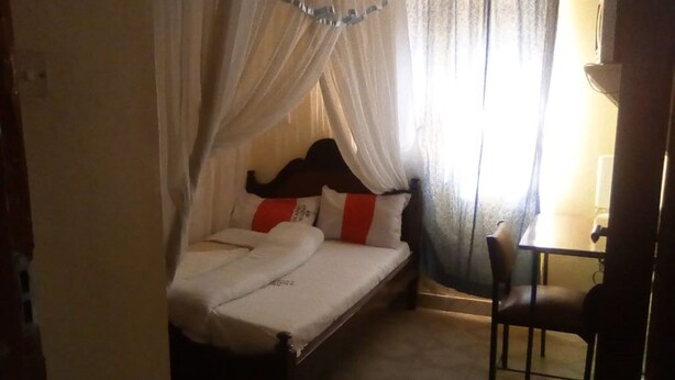 A single bed with a canopy, a bedside table, and a chair in a room with a tiled floor. - Scarlet Holiday Inn (Nakuru)